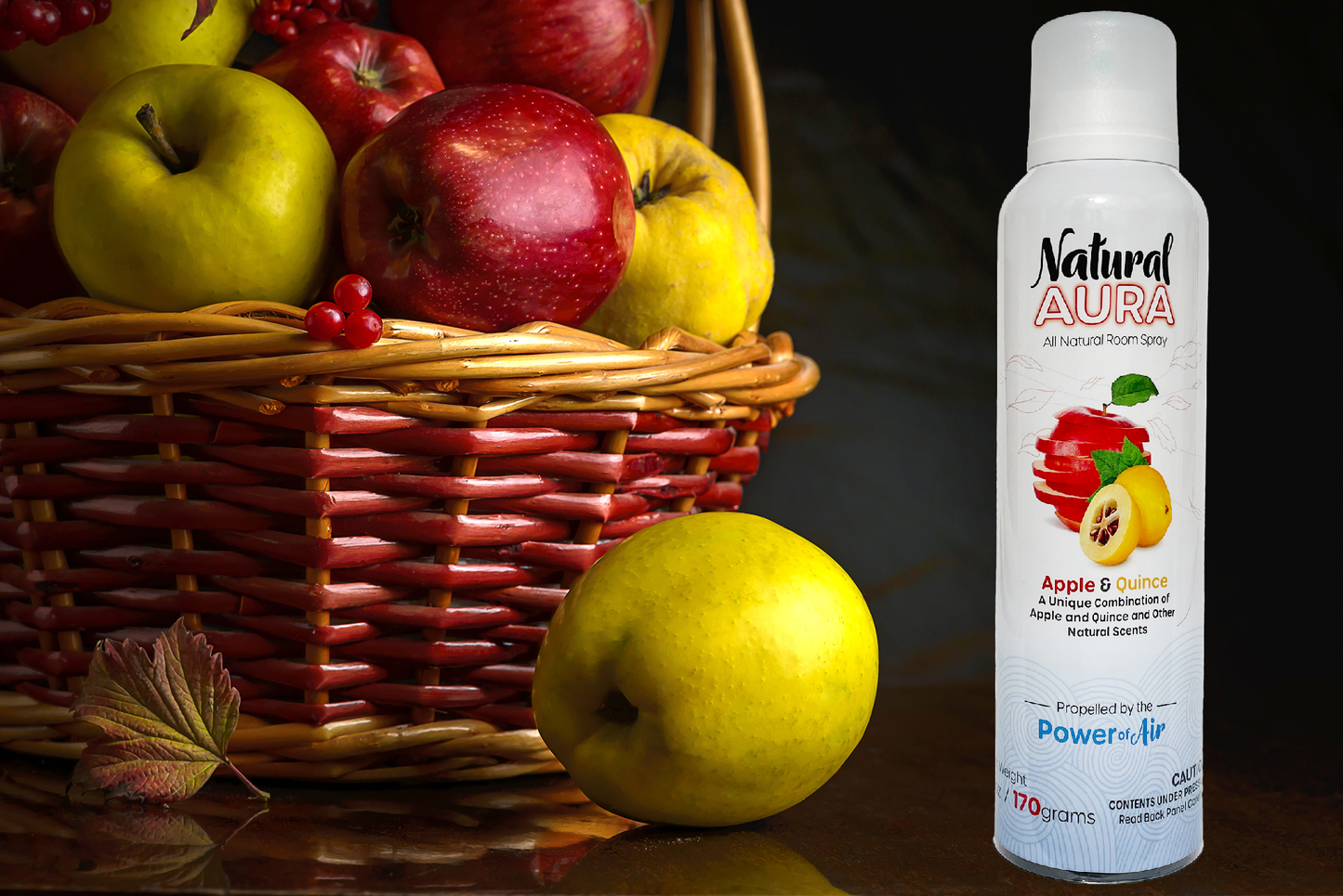Bottle of Natural Aura Apple and Quince Room Spray sitting on a dark brown table next to a wicker basket of red apples and yellow quince.