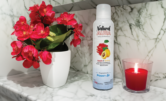 Natural Aura Apple and Quince Room Spray sitting on a bathroom white and black marble counter with red flowers and a red lit candle.