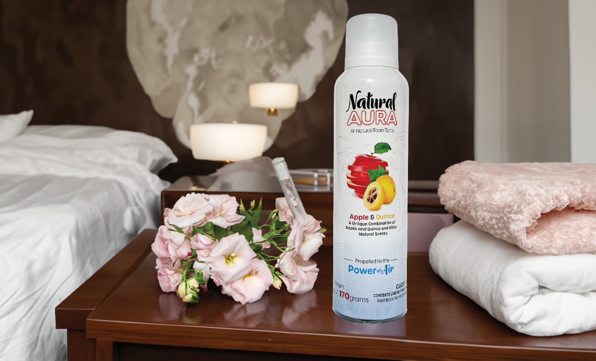 Bottle of Natural Aura Apple and Quince room spray sitting on a brown wooden end table with a small pink bouquet of flowers and a pink towel and white towel all in a bedroom with a bed in the background.