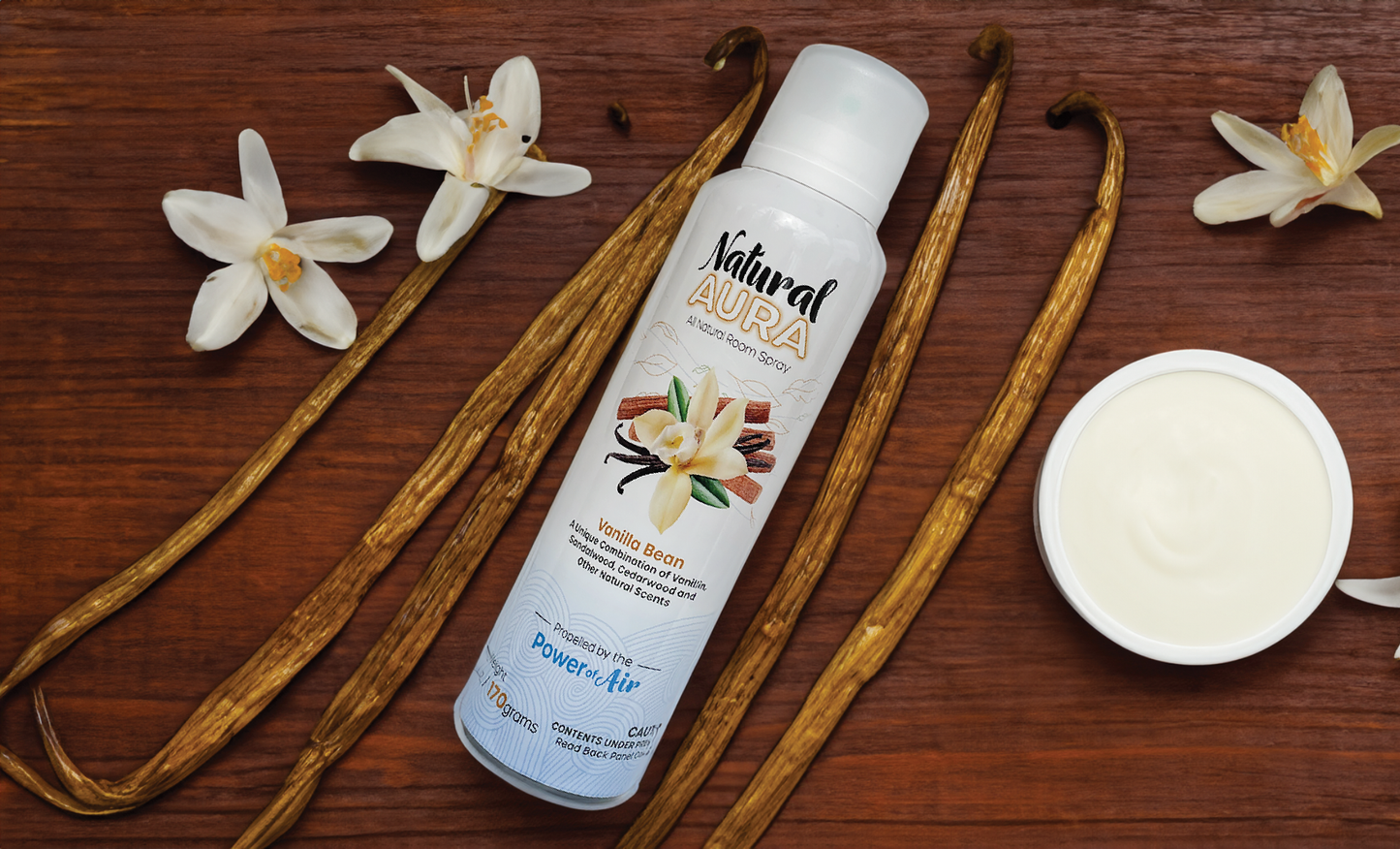 Bottle of Natural Aura Vanilla Bean Room Spray laying down on a brown wooden table with vanilla bean sticks and vanilla flowers.