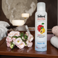 Bottle of Natural Aura Apple and Quince room spray sitting on a brown wooden end table with a small pink bouquet of flowers and a pink towel and white towel all in a bedroom with a bed in the background.