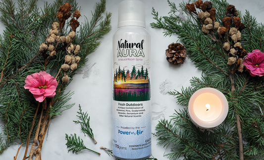 Bottle of Natural Aura Fresh Outdoors Room Spray on a white table with forest green pine branches and a pine cone and red flowers and a lit candle.