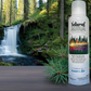 bottle of Natural Aura Fresh Outdoors room spray with a waterfall cutting through the forest with pine trees and soft velvety moss in the background.