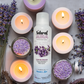 bottle of Natural Aura Lavender Breeze Room Spray laying down on a table surrounded by white and purple candles that are lit as well as lavender flowers laying on the table.
