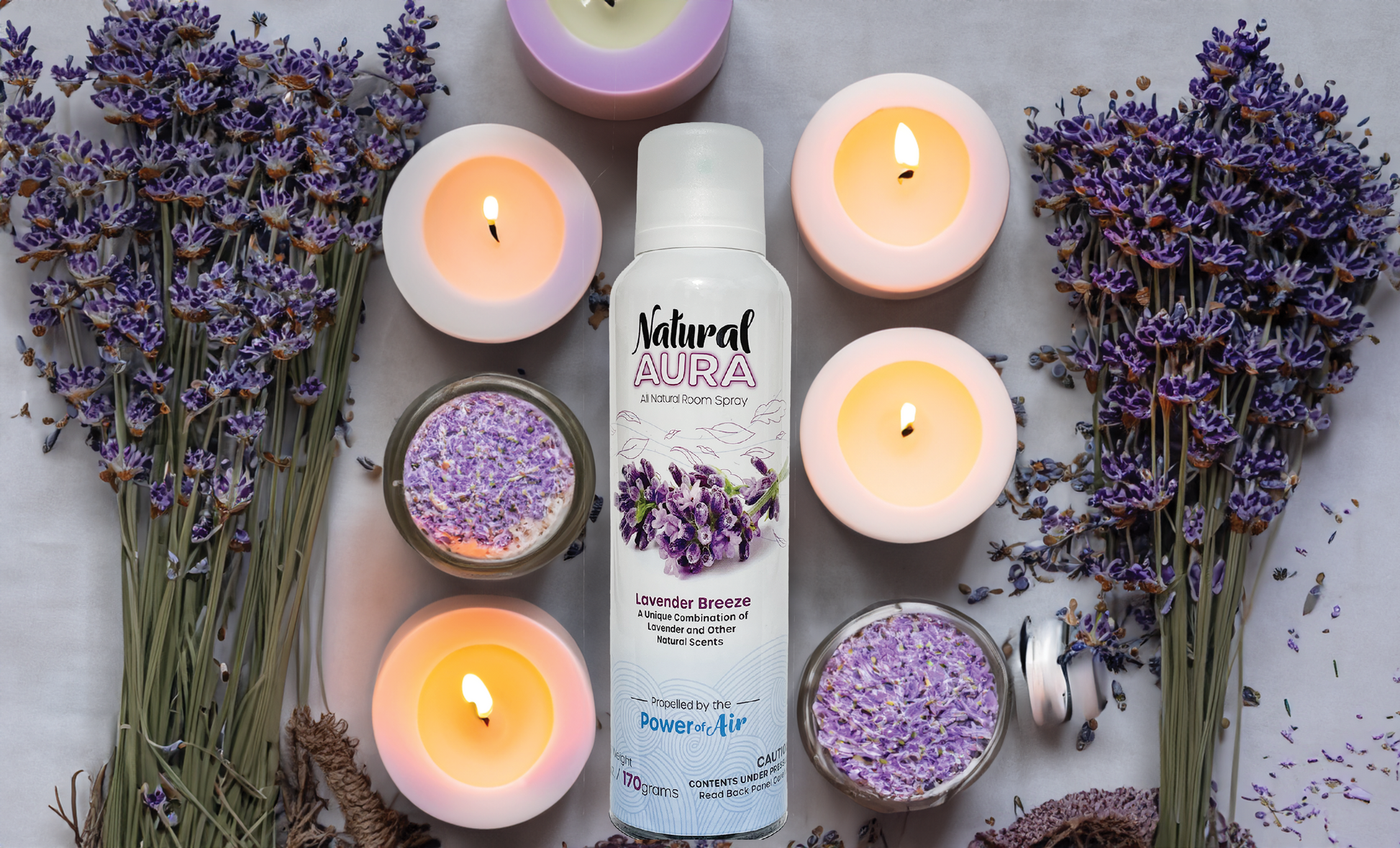 bottle of Natural Aura Lavender Breeze Room Spray laying down on a table surrounded by white and purple candles that are lit as well as lavender flowers laying on the table.