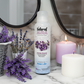 Bottle of Natural Aura Lavender Breeze Room Spray sitting on a white and grey marble bathroom countertop with 2 round mirrors in the background along with white and purple candles as well as lavender in a basket and loose stems of lavender on the countertop.