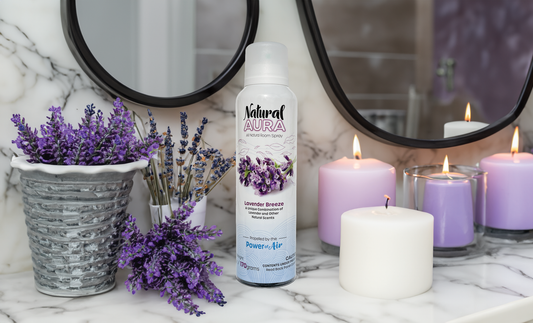 Bottle of Natural Aura Lavender Breeze Room Spray sitting on a white and grey marble bathroom countertop with 2 round mirrors in the background along with white and purple candles as well as lavender in a basket and loose stems of lavender on the countertop.