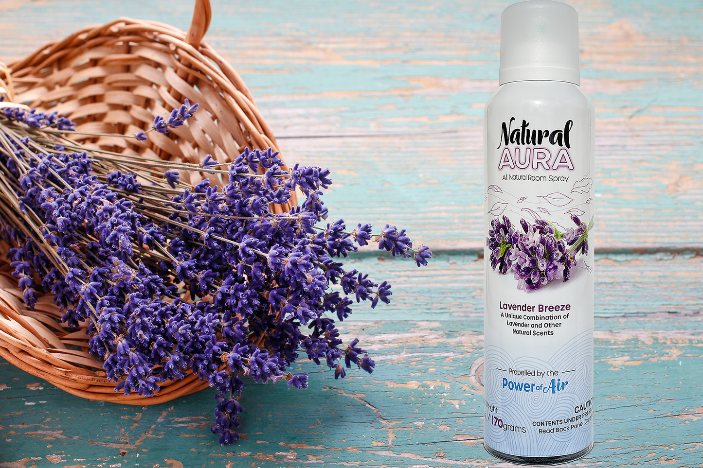 Bottle of Natura Aura Lavender Breeze Room Spray sitting on a wooden table with cracking teal paint along with a wicker basket containing purple lavender branches.