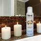 Natural Aura Vanilla Bean Room Spray sitting on a brown marble bathroom counter along with white candles that are lit with a mirror in the background.