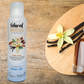 Bottle of Natural Aura Vanilla Bean Room Spray on a tan wooden table with vanilla bean sticks and vanilla extract oil in a clear glass bottle with a cork top.